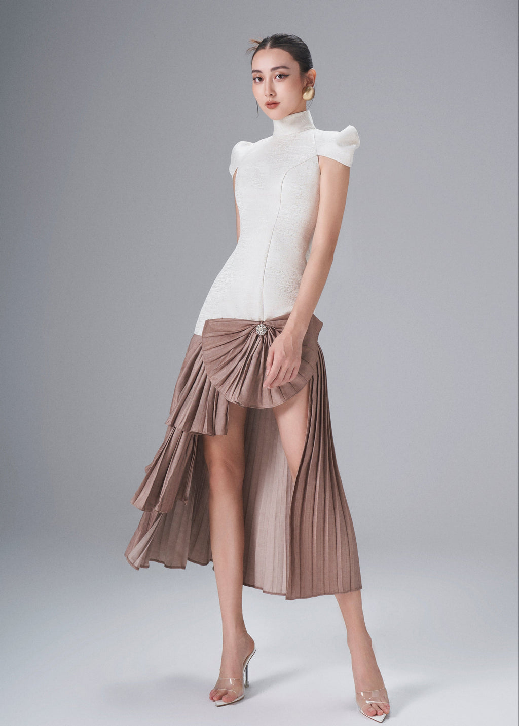 Woman wearing a white top and brown pleated skirt on a gray background